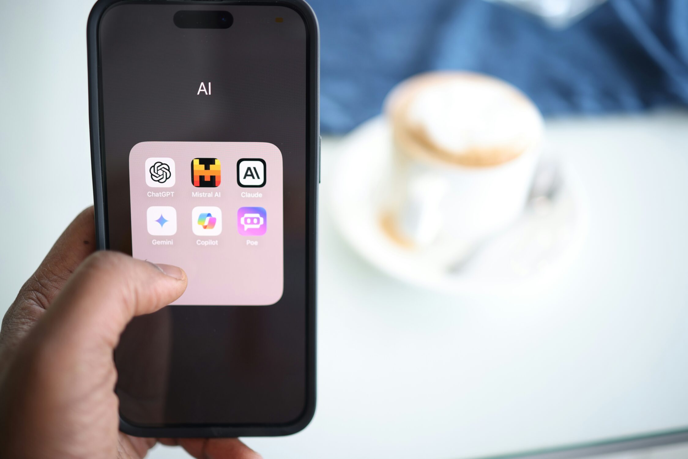 A hand holding a phone showing a folder of AI apps including ChatGPT, Claude and Gemini